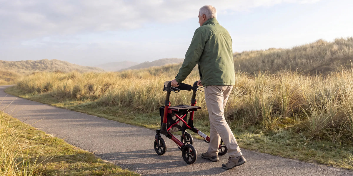 Parvum Brava in ruby red on a dune path in the morning mist; a man walks with the rollator.
